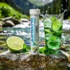 ÉLECTROLYTES en bouteille blanche sur roche près d’une rivière, verre de boisson pétillante verte avec rondelles de citron vert et feuilles de menthe, eau et splash derrière.